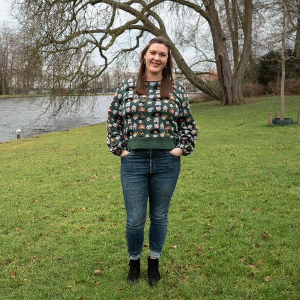 Brianna stands on the park lawn wearing black boots, blue jeans and a dark green, home-sewn  sweater with faces of multi ethnic ladies on it. In the background, a large oak tree dips into the water.