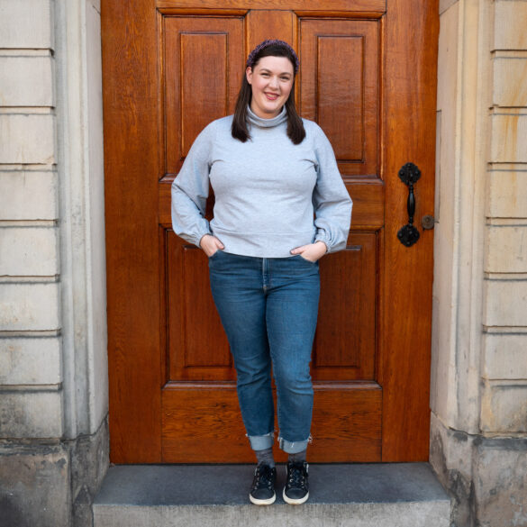 A white woman stands in front of a church door smiling. She's wearing a gray turtleneck sweater.
