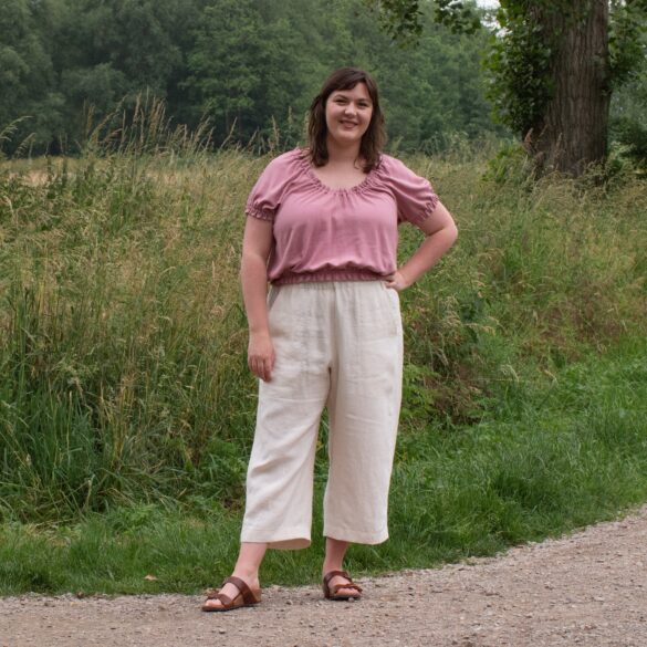 Bri, a white femme, stands by a field wearing tan wide leg pants and a pink peasant style crop top.